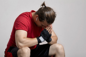 Young caucasian man in sport wear and boxing bandages sits with hands to forehead. Pray, or despair moment, sorrow, hopelessness, despond before or after match. Indoors, copy space, light background.