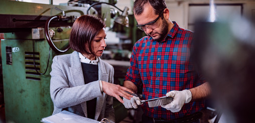 Machinist standing with female engineer and measuring cogwheel diameter
