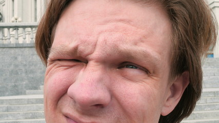 Obraz premium portrait of a white charismatic man looking at the sun and camera on city architecture background and squinting laughing slow motion close up video in 4K