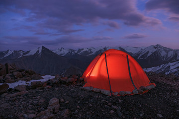 Safe travel concept: tent set in the high mountains is reinforced around the perimeter with stones for stability in conditions of strong wind. Tian Shan peaks, twilight, last light after sunset.