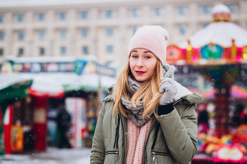 A girl in a pink hat and a knitted woolen scarf walks at the Christmas market. blonde girl smiles and laughs on the background of the winter fair.