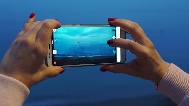 A woman holds a smartphone in her hands and takes pictures of the sea after sunset