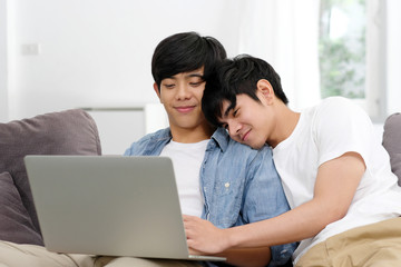 Happy young asian gay man couple using laptop computer while sitting onsofa at home, homosexual and lgbt with technology lifestyle concept