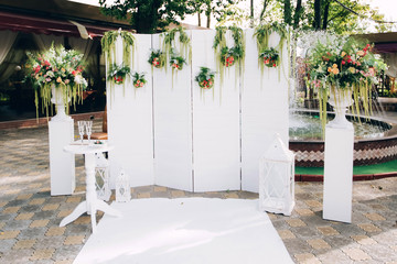 beautiful wedding photo zone in the park with fountains in the shade, a white screen, a pedestal with flowers and decor. Wedding screen
