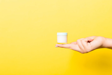Female hand holding cream bottle of lotion isolated. Girl give jar cosmetic products on yellow background