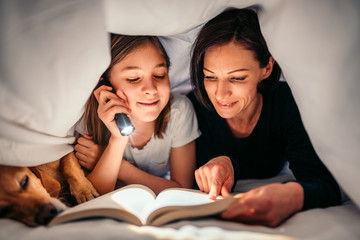 Mother, daughter and dog laying on the bed and reading book late at night
