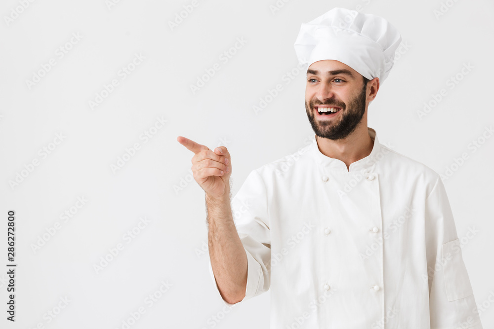 Wall mural cheery optimistic young chef posing isolated over white wall background in uniform pointing aside.