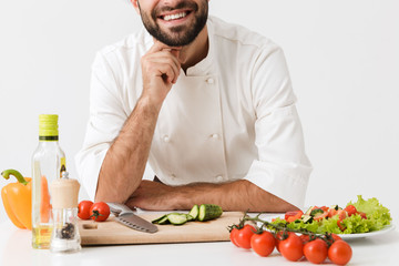 Pleased positive happy young chef isolated over white wall background in uniform cooking with fresh vegetables.
