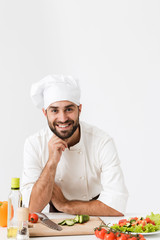 Pleased positive happy young chef isolated over white wall background in uniform cooking with fresh vegetables.