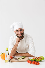 Pleased positive happy young chef isolated over white wall background in uniform cooking with fresh vegetables.