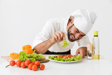 Pleased positive happy young chef isolated over white wall background in uniform cooking with fresh vegetables.