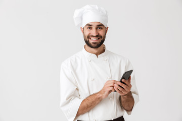 Cheerful smiling young chef posing isolated over white wall background in uniform using mobile phone.