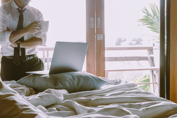 Businessman holding cup of coffee and working on laptop in bed room.