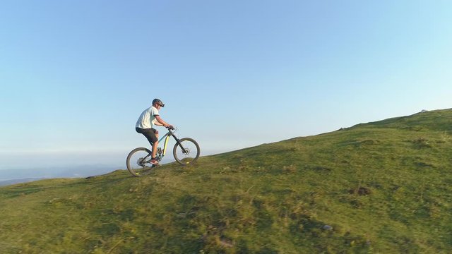 DRONE: Flying along an athletic man pedalling an e-bike up a steep grassy hill. Male tourist downhill biking in the Slovenian mountains riding his electric bicycle uphill on a sunny summer day.
