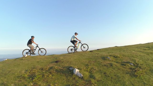 DRONE: Two mountain bikers ride their electric bicycles up a steep grassy hill before descending into the green valley in the picturesque Slovenian countryside. Fit male tourists cruising on e-bikes.
