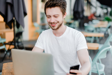 young man doing freelance work with phone and laptop in cafe outdoors