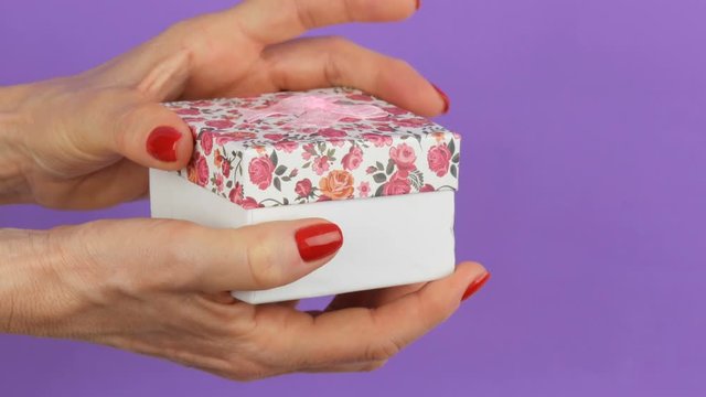 Open a small gift box with a floral print and a pink bow that is held by the hands of a woman whose nails are painted with red nail polish on a purple background.