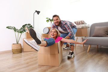 Young married couple moving into a new home. Attractive blonde woman sitting in cardboard box, bearded man pushes her. Newely weds fooling around. Minimal interior background, copy space, close up