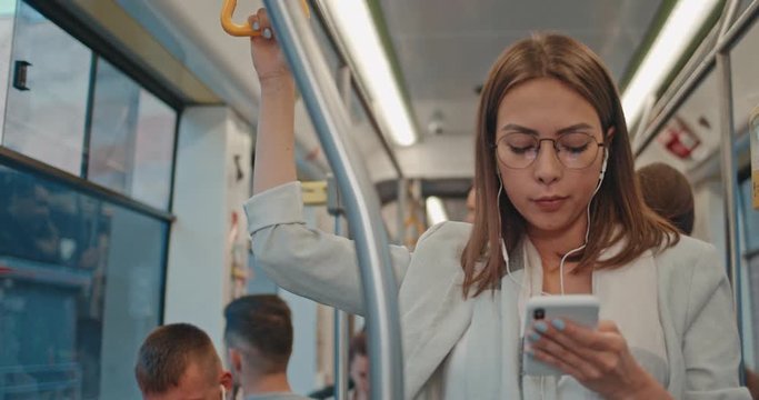 Portrait of serious girl wearing glasses and earphones holds the handrail and using smartphone, listening music. Attractive women browsing on mobile phone in public transport. City, urban background.