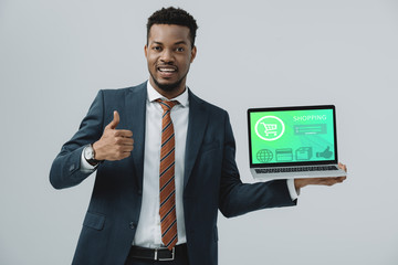 cheerful african american man holding laptop with shopping website on screen and showing thumb up isolated on grey