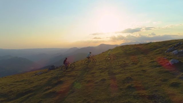 AERIAL, COPY SPACE, SUN FLARE: Flying along three active tourists riding their electric bicycles up a grassy hill on a beautiful summer morning. Cinematic shot of young tourists on mountain bike ride.