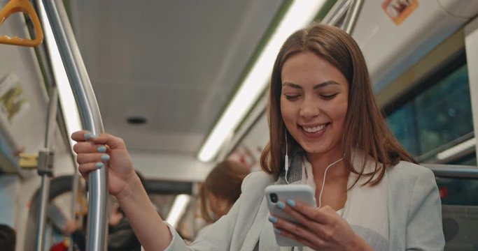 Portrait of smilling girl wearing earphones holds the handrail and using smartphone, listening music. Attractive women browsing on mobile phone in public transport. People on background.