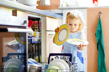 Little adorable cute toddler girl helping to unload dishwasher. Funny happy child standing in the...