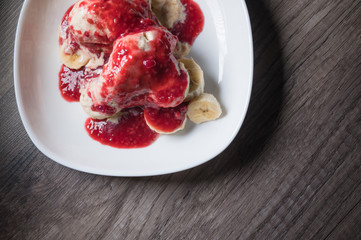 Closeup Vanilla ice cream poured with strawberry or raspberry jam with sliced banana slices on a white plate on a dark wooden table. You can do this at home on vacation