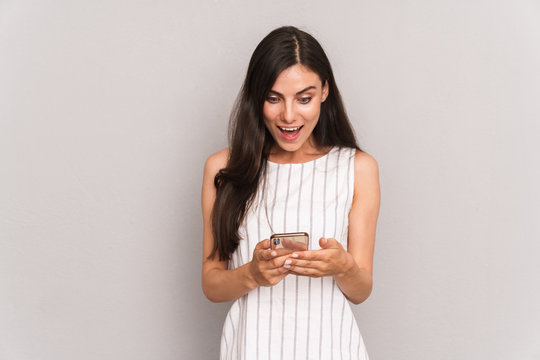 Image of pretty brunette woman wearing dress smiling while holding modern smartphone