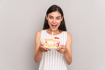 Image of happy brunette woman wearing dress smiling and holding birthday cake with candle