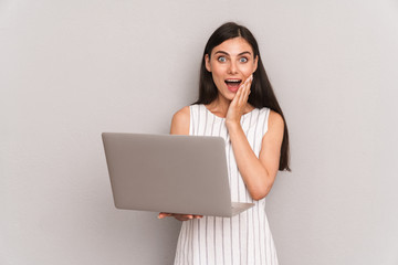 Image of excited brunette woman wearing dress smiling while holding silver laptop computer