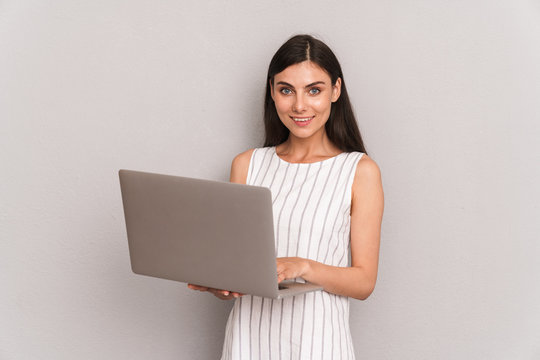 Image of attractive brunette woman wearing dress smiling while holding silver laptop computer