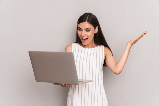 Image of emotional brunette woman wearing dress smiling while holding silver laptop computer