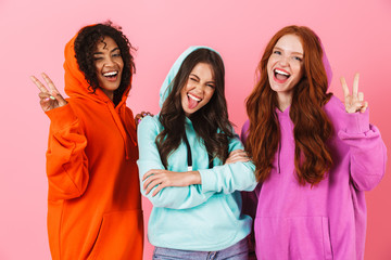 Pleased smiling young three multiethnic girls friends posing isolated over pink wall background...