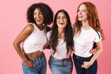 Happy young three multiethnic girls friends posing isolated over pink wall background.