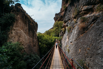 view of a woman with a leather backpack crossing a hanging bridge into the forest.