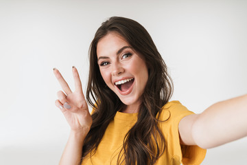 Image of pleased brunette woman smiling and showing peace sign while taking selfie photo