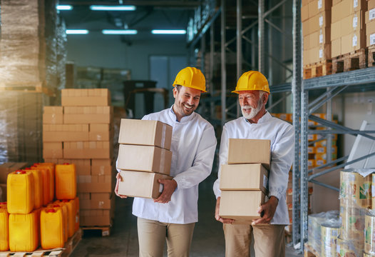 Three quarter length of two smiling Caucasian storage employees in white uniforms and with yellow helmets carrying boxes in storage.