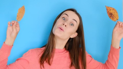 Portrait of young girl with long hair wearing pink sweater, having fun playing with yellow autumn leaves and looking at camera, against blue background. People and lifestyle.