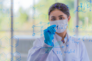 A young beautiful asian girl scientist wear health mask writing table of the elements on glass board in laboratory at morning classroom. Preparing for new biologic lab. Selective focus on woman face.