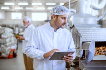 Young Caucasian serious supervisor evaluating quality of food in food plant while holding tablet. Man is dressed in white uniform and having hair net.