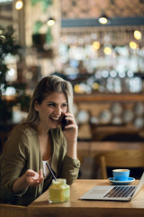 Happy young woman talking on smart phone while working on laptop in a cafe