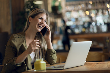 Happy young businesswoman talking on smart phone while working on laptop in a cafe