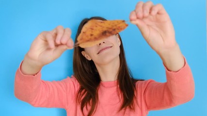 Portrait of young girl with long hair wearing pink sweater, having fun playing with yellow autumn leaves and looking at camera, against blue background. People and lifestyle.