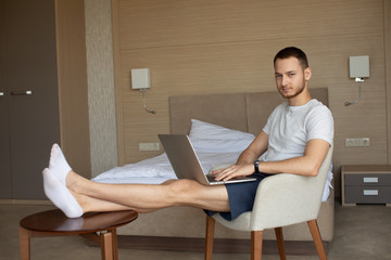 Smiling young man is working with laptop, sitting on armchair.