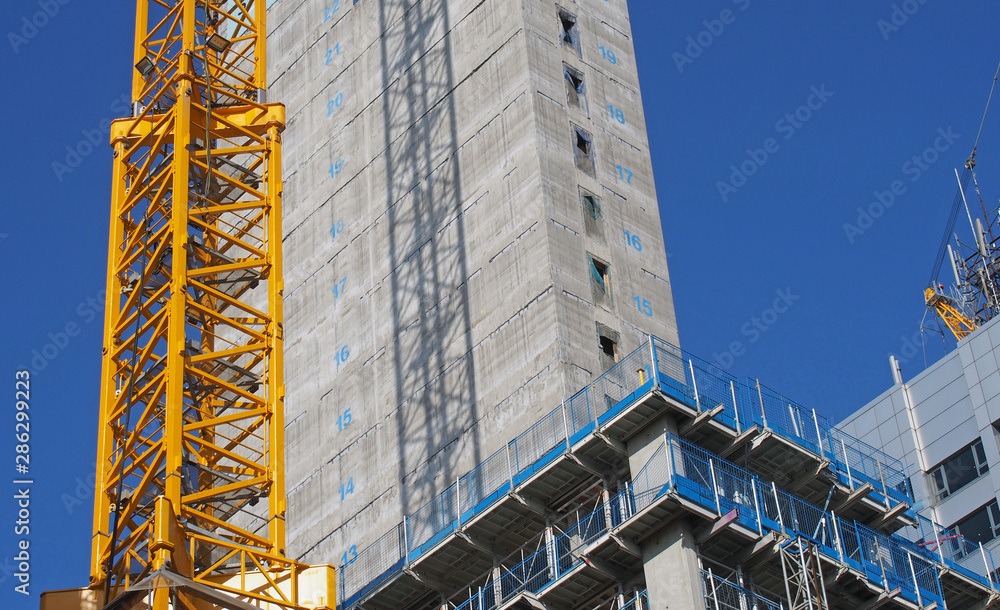 Wall mural close up of a large urban construction site with a yellow tower crane casting a shadow on a large concrete building and safety fences against a blue sky