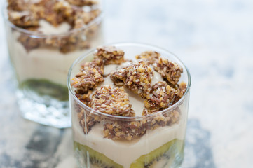 Granola, kiwi, banana and Greek yogurt in two glass cups on gray concrete, close-up macro