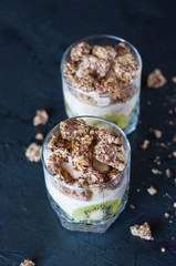 Greek yogurt, granola, banana and kiwi. Desserts in glass cups on dark background, vertical format. Concept body and healthy food