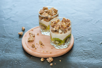 Greek yogurt, granola, banana and kiwi. Desserts in glass cups with spoons on a wooden round shelf, dark background, side view. Concept body and healthy food
