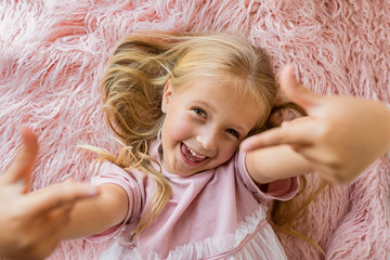 Adorable little girl with long blonde hair lying on pink fur blanket and making selfie. Positive emotions , expression, fun. Happy smiling kid, childhood day, education, kids using technology concept
